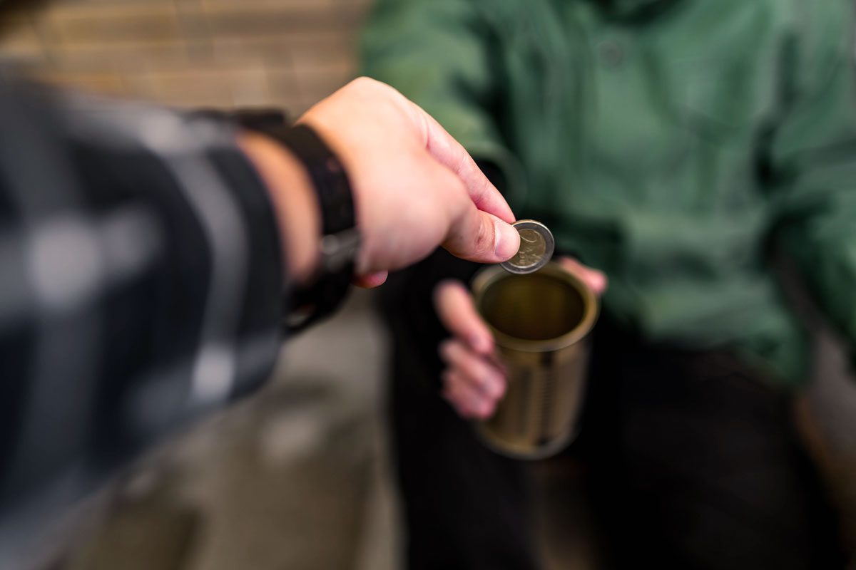 A close-up of a hand dropping a coin into a tin can held by a person wearing a green jacket.