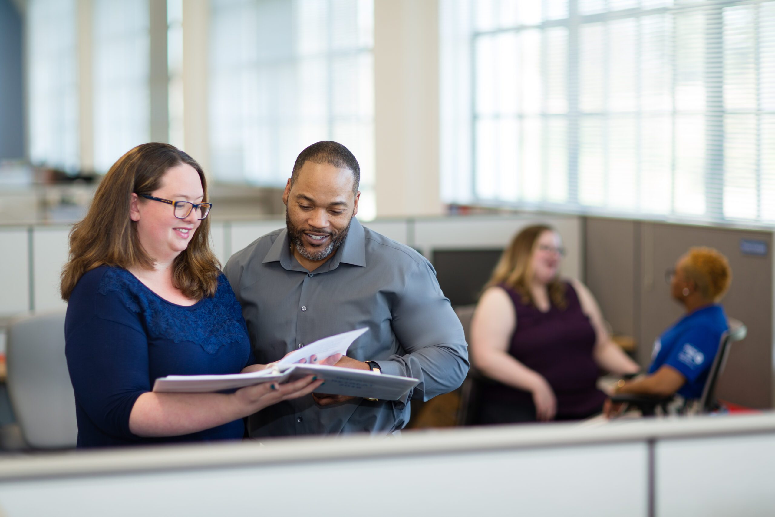 A woman in a navy blue blouse and a man in a gray shirt review a document together in an office setting, both smiling. In the background, two other colleagues are engaged in conversation near cubicles with large windows letting in natural light.