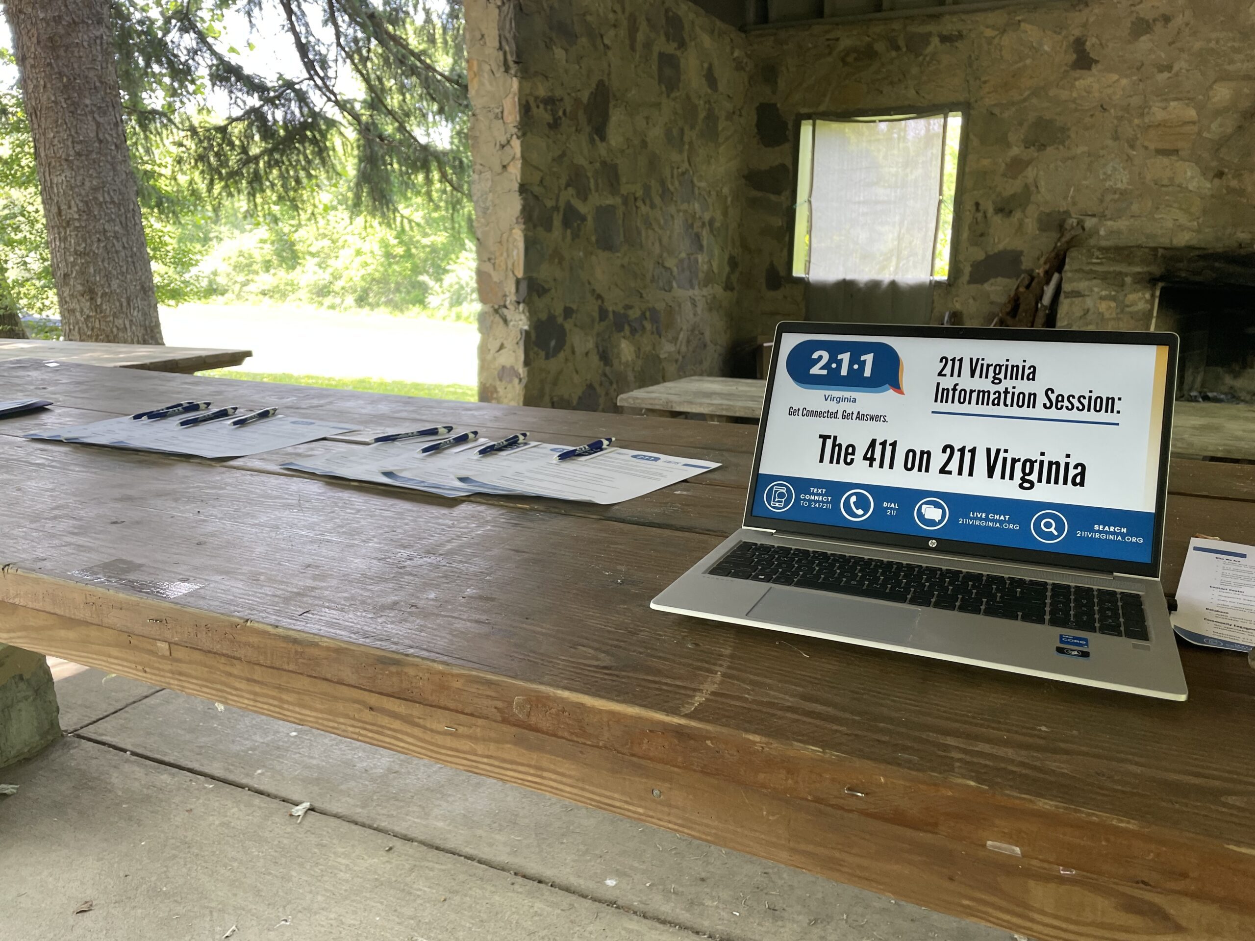 A wooden picnic table holds sign-up sheets with pens and an open laptop displaying a “211 Virginia Information Session” presentation. The setting is an outdoor pavilion with a stone wall, a window with a curtain, and a fireplace in the background.