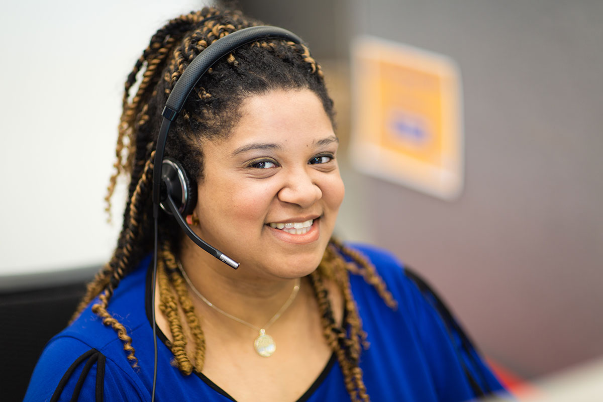 A smiling woman wearing a headset and a blue top sits at a workstation in a call center.