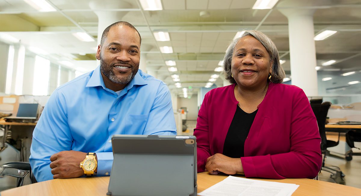 A man in a light blue shirt and a woman in a red cardigan sit together at a table, smiling while looking at a tablet. The setting is a bright office with large windows and cubicles in the background.