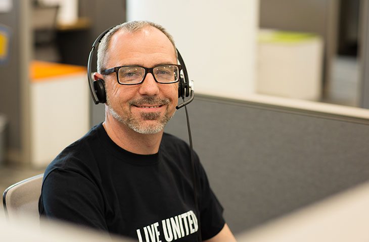 A smiling man wearing glasses and a headset sits at a workstation in a call center. The background features office cubicles with a neutral color scheme.