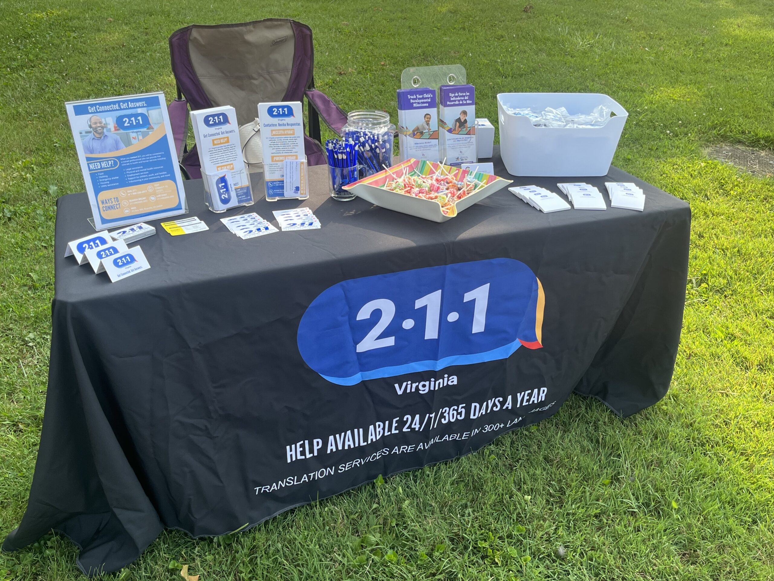 An outdoor informational booth for 211 Virginia features brochures, flyers, pens, business cards, and a tray of candy on a table covered with a branded tablecloth. A folding chair is set up behind the table, which is placed on a grassy area.