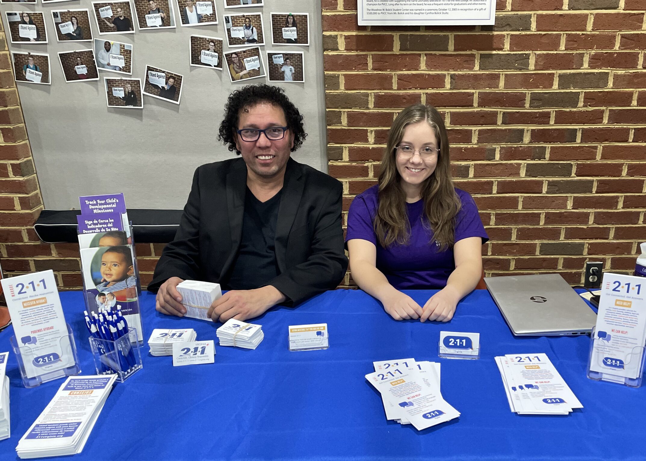 Two smiling representatives sit behind a table covered with a blue cloth, displaying brochures, pens, and informational materials about 211 services. A brick wall with pinned photos and a laptop are visible in the background.