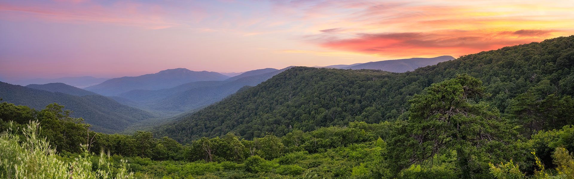 A lush, green mountain landscape stretches into the distance under a vibrant sunset sky with shades of orange, pink, and purple. Layers of rolling hills and valleys create depth, with dense forests covering the slopes.