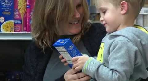 A smiling woman holds a young boy in her arms as they look at a box of macaroni and cheese in a grocery store aisle. Shelves stocked with pasta products are visible in the background.