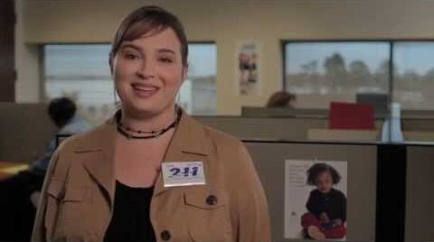 A woman wearing a tan jacket and a “211” name tag smiles while standing in an office with cubicles. In the background, employees are working at their desks.