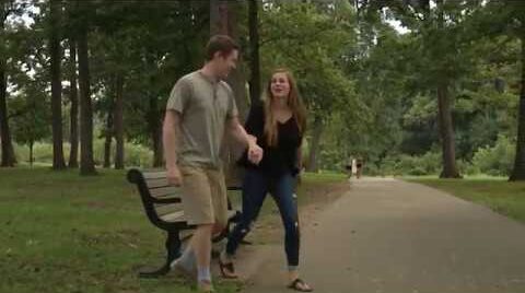 A young man and woman walk along a paved path in a park, holding hands.