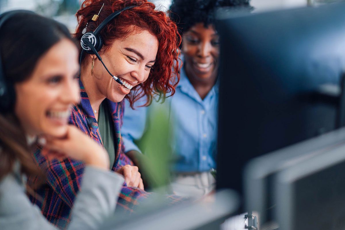 Three smiling women wearing headsets work together at a computer in a call center. They appear engaged and happy as they assist callers or collaborate on a task.